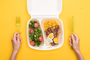 cropped view of woman holding plastic fork, knife and  package with corn, meat, fried eggs and salad isolated on yellow