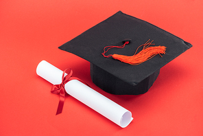Academic cap with tassel and diploma with ribbon on red surface
