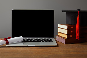 Laptop with blank screen, books, diploma and academic cap on wooden table isolated on grey