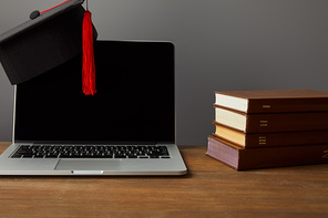 Laptop with blank screen, books and academic cap with red tassel on wooden surface isolated on grey