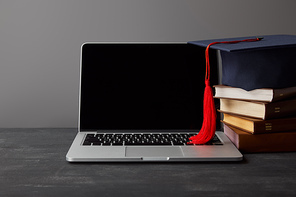 Laptop with blank screen, books and academic cap with red tassel on grey
