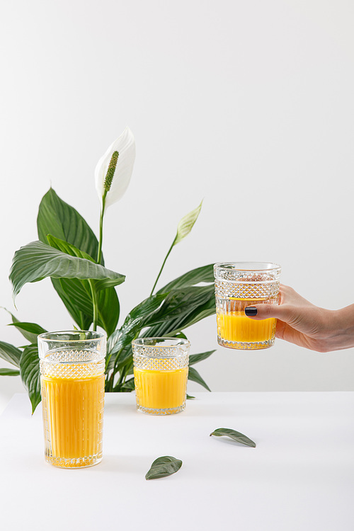 cropped view of woman holding glass of fresh delicious yellow smoothie near green peace lily plant isolated on white