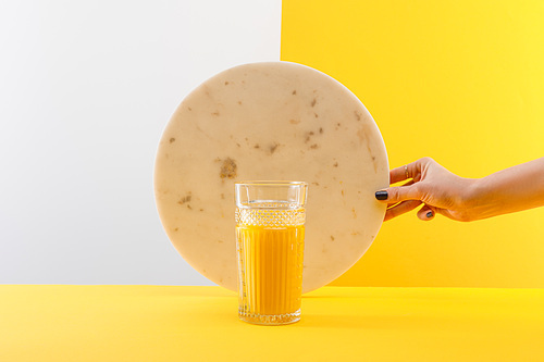 cropped view of woman holding marble circle near glass of fresh delicious yellow smoothie on grey and yellow background