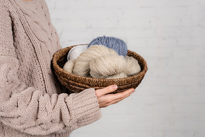 Cropped view of woman in knitted sweater holding basket with woolen yarn on white background