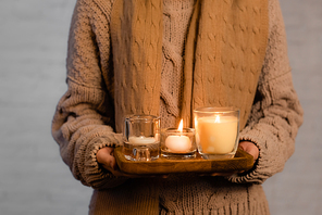 Cropped view of burning candles on wooden board in hands of woman in scarf and sweater blurred on white background