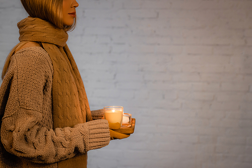 Cropped view of woman in sweater, scarf and gloves holding burning candles near white brick wall