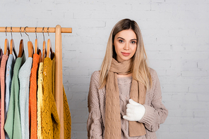 Smiling woman in knitwear and glove standing near hanger rack with sweaters on white background