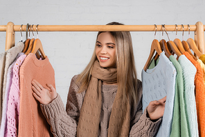 Cheerful woman in scarf looking at sweaters on hanger rack on white background