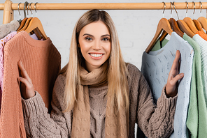 Woman in scarf smiling at camera near warm sweaters on hanger rack on white background