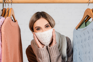 Woman covering face with scarf near sweaters on hanger rack on white background