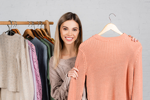 Positive woman holding cozy sweater near knitwear on hanger rack on blurred on white background