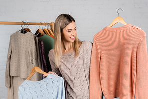 Smiling woman holding hangers with sweaters near hanger rack blurred on white background