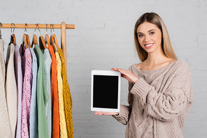 Smiling woman holding digital tablet with blank screen near hanger rack with sweaters on white background