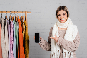Smiling woman pointing at smartphone with blank screen near hanger rack with sweaters on white background