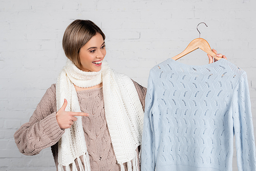 Cheerful woman in scarf pointing at hanger with sweater on white background