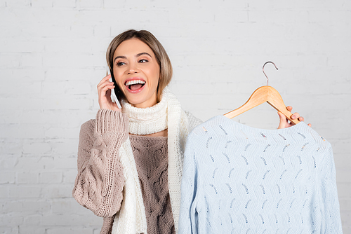 Cheerful woman in warm clothes talking on cellphone and holding hanger with sweater on white background