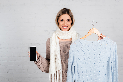 Young woman in scarf holding smartphone with blank screen and sweater on hanger on white background
