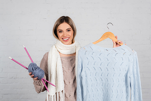Smiling woman holding hanger with sweater and yarn with knitting needles on white background