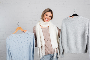 Smiling woman in scarf holding sweaters on hangers on white background