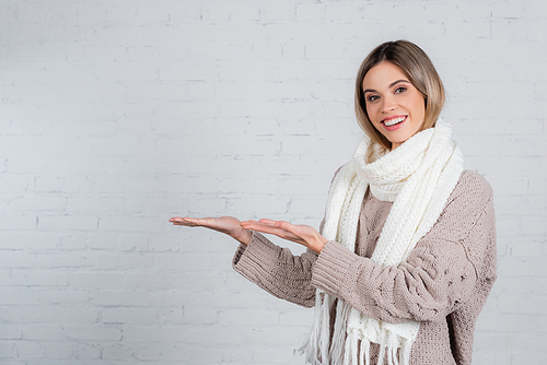 Smiling woman in scarf and sweater pointing with hands near white brick wall