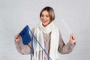 Positive woman in scarf and sweater holding yarn and knitting needles near on white brick wall