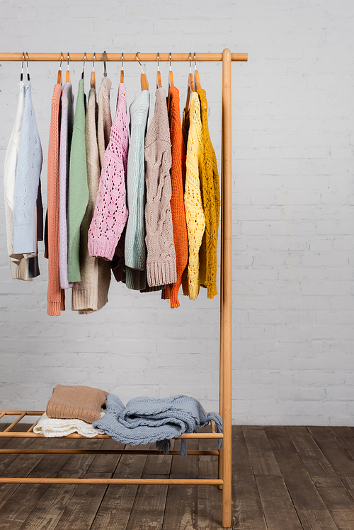 Wooden hanger rack with colorful knitted sweaters near white brick wall