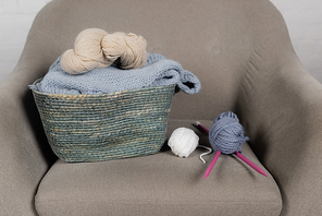 Basket with woolen yarn and knitting needles on armchair on white background