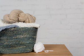 Basket with woolen yarns on wooden table near white brick wall