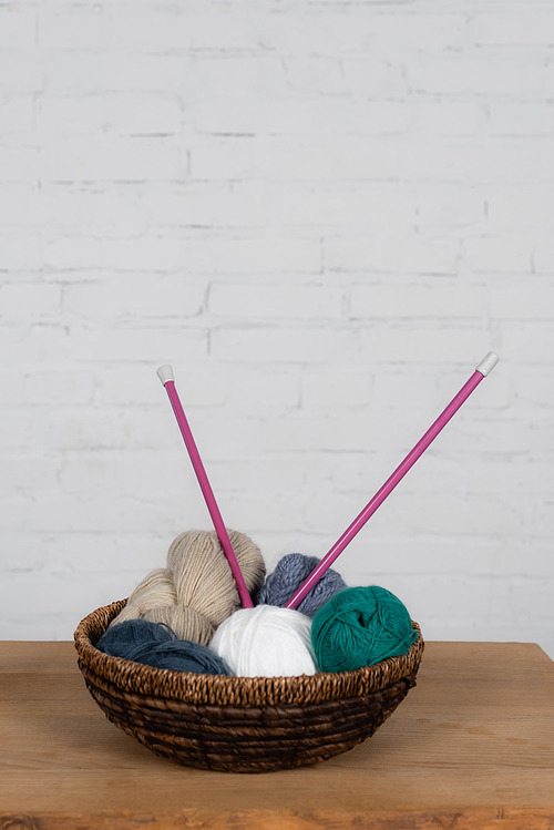 Needless in basket with yarn balls on wooden table and brick wall at background