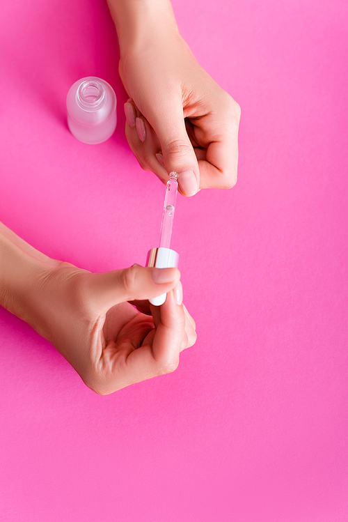 partial view of woman applying cuticle remover with dropper on pink background