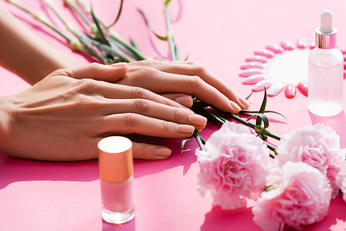 cropped view of female hands near carnation flowers, bottles of nail polish and cuticle remover, and fake nails palette on pink