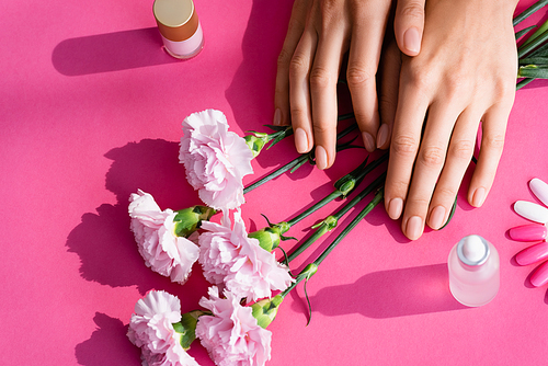 top view of female hands near carnation flowers, palette of fake nails, vials with cuticle remover and nail varnish on pink background