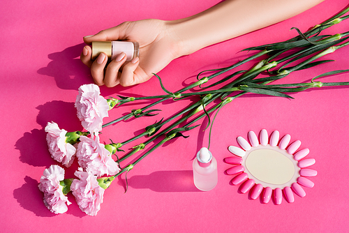 cropped view of woman holding pastel nail polish near carnation flowers, cuticle remover and palette of artificial nails on pink background