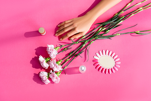 top view of carnation flowers near female hand, palette of artificial nails, nail polish and cuticle remover on pink background