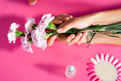 cropped view of woman holding carnation flowers near palette of artificial nails and cuticle remover on pink background