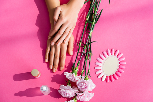 top view of female hands near palette of fake nails, carnation flowers, cuticle remover and nail varnish on pink background