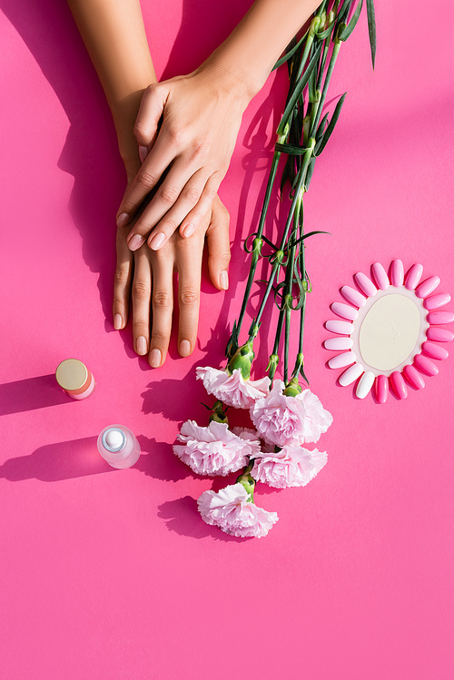 top view of female hands near carnation flowers, palette of artificial nails, cuticle remover and nail polish on pink background