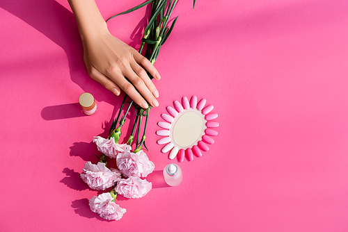 top view of female hands near bottles with nail varnish and cuticle remover, carnation flowers and palette of fake nails on pink background