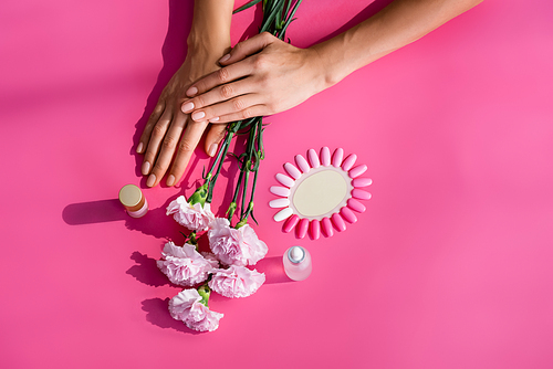 top view of female hands near palette of false nails, carnation flowers, and bottles of cuticle remover and enamel on pink background