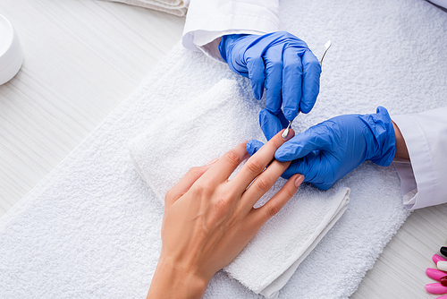 partial view of manicurist in latex gloves making manicure to woman with cuticle pusher