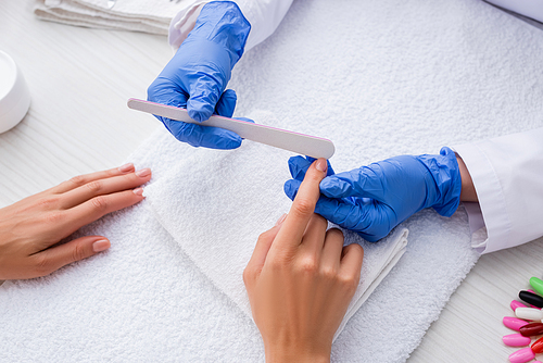 partial view of manicurist in latex gloves using nail file while making manicure to woman