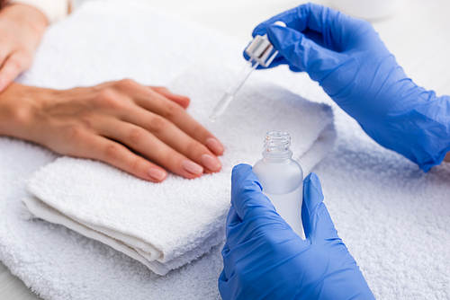 partial view of manicurist in latex gloves applying cuticle remover on finger of client, blurred background