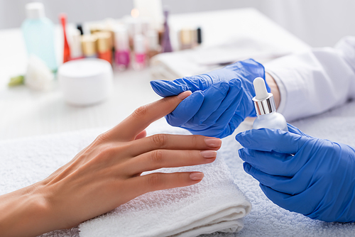 partial view of manicurist holding vial of cuticle remover while touching finger of client, blurred background