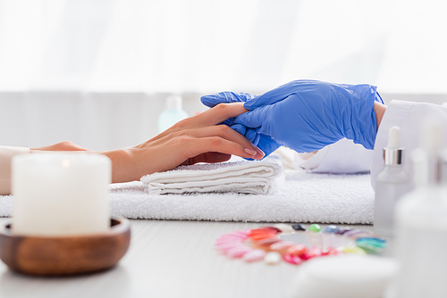 partial view of manicurist in latex gloves touching hand of client on blurred foreground