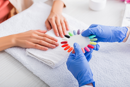partial view of manicurist showing palette of fake nails to client on blurred background