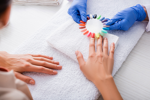 partial view of manicurist in latex gloves holding palette of false nails near hand of woman