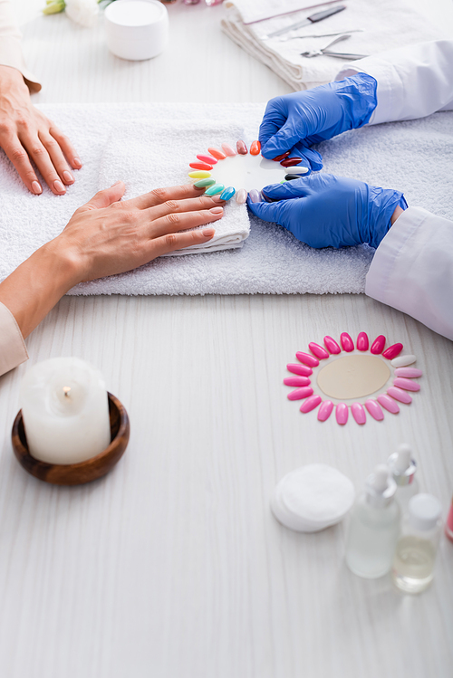 cropped view of manicurist holding set of artificial nails near client and manicure supplies on blurred foreground