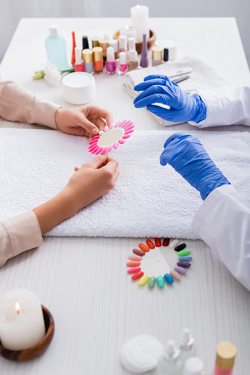 cropped view of woman holding palette of fake nails near manicurist and manicure supplies on blurred background