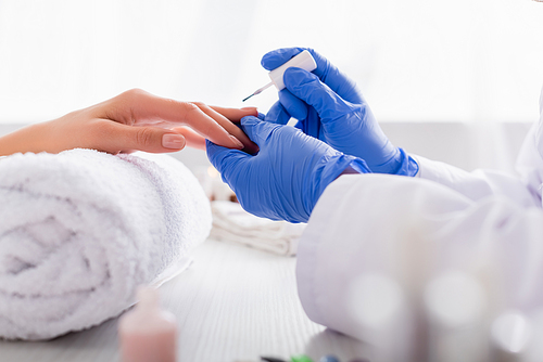 partial view of manicurist in latex gloves applying nail polish while making manicure to client on blurred foreground