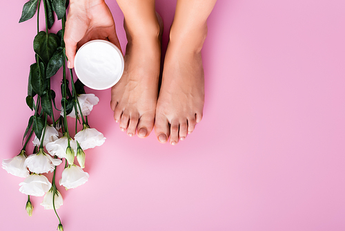 cropped view of woman holding cosmetic cream near groomed feet and white eustoma flowers on pink background